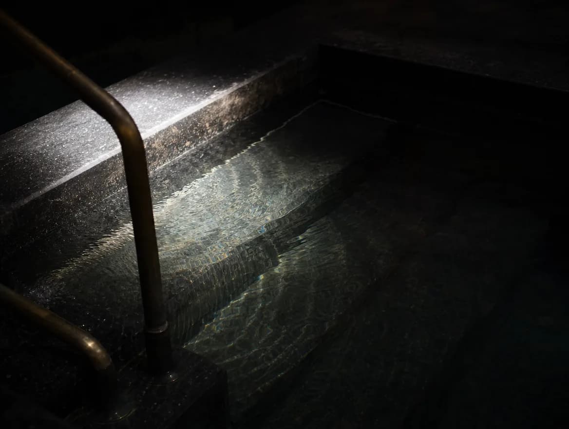 The surface of a dark stone cold plunge with an aged bronze step rail.